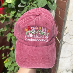 May include: A maroon baseball cap with embroidered flowers in various colors. The text "radicalized by basic decency" is embroidered below the flowers. The cap has a curved brim and a fabric strap closure.