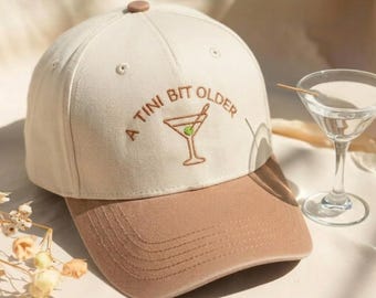 May include: A cream-colored baseball cap with a brown brim and a brown button on top. The front features brown embroidered text that reads A TINI BIT OLDER above an embroidered martini glass graphic with a small green olive inside the glass.