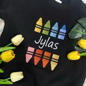 Op de afbeelding: Zwart sweatshirt met een kleurpotloontje. Het ontwerp bevat vier kleurpotloden in geel, groen, blauw en lichtblauw boven de naam "Jylas" in witte letters. Daaronder staan nog vier kleurpotloden in roze, oranje, rood-oranje en geel-oranje.