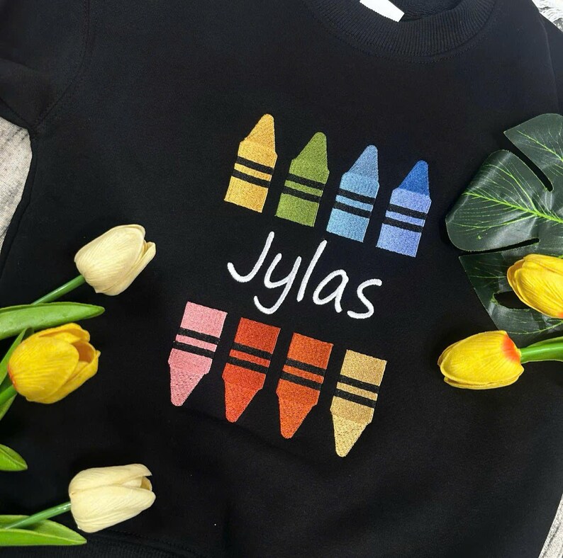 Op de afbeelding: Zwart sweatshirt met een kleurpotloontje. Het ontwerp bevat vier kleurpotloden in geel, groen, blauw en lichtblauw boven de naam "Jylas" in witte letters. Daaronder staan nog vier kleurpotloden in roze, oranje, rood-oranje en geel-oranje.