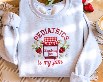 May include: Light gray sweatshirt with the embroidered text "PEDIATRICS" above a strawberry jam jar illustration. The jar is surrounded by green leaves and red strawberries, with the phrase "is my jam" below.
