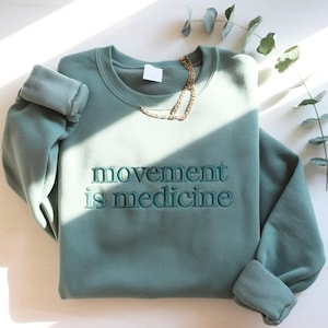 May include: A teal-colored sweatshirt with the embroidered text "movement is medicine" in a matching teal thread. A gold chain necklace rests on the sweatshirt. The sleeves are cuffed, and the garment is displayed on a white surface.