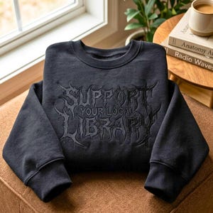 Peut inclure: Un sweat-shirt à col rond gris anthracite avec une inscription brodée noire SUPPORT YOUR LOCAL LIBRARY dans une police gothique stylisée et pointue. Le vêtement possède des manches longues avec des poignets et un col côtelés.