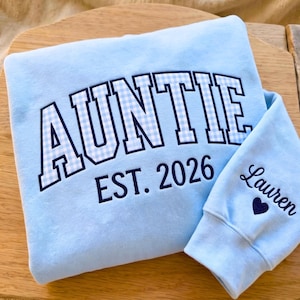 May include: Light blue sweatshirt with the word "AUNTIE" in large, white, and blue gingham letters. The text "EST. 2026" is below. A matching sleeve has the name "Lauren" and a heart. The sweatshirt is on a wooden surface.