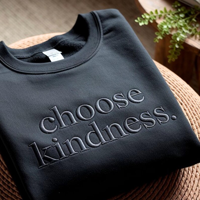 May include: A black crewneck sweatshirt with the embroidered phrase "choose kindness." The sweatshirt is made of a soft material and is folded neatly. The text is in a simple, elegant font.