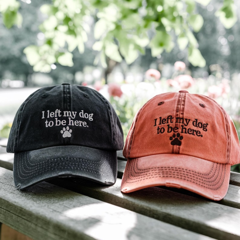 May include: Two distressed baseball caps with the text "I left my dog to be here." and a paw print. One cap is black and the other is orange.