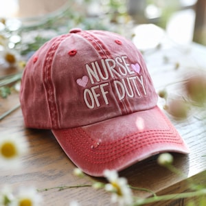 May include: Red distressed baseball cap with white embroidery that reads "Nurse Off Duty" with two pink hearts.