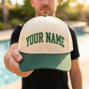 May include: A beige baseball cap with a dark green brim and the text "YOUR NAME" embroidered in dark green capital letters across the front panel. The cap features a structured crown and a button on top, showcasing a two-tone color scheme.