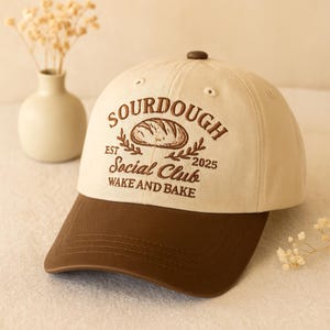 May include: A beige and brown baseball cap with the text "SOURDOUGH Social Club WAKE AND BAKE" embroidered on the front. The cap features a brown brim and a brown button on top. The design includes an illustration of a loaf of bread.