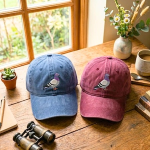 Puede incluir: Dos gorras de béisbol, una azul y otra burdeos, están sobre una superficie de madera. Cada gorra presenta un diseño de paloma bordada. Las gorras están cerca de una ventana, una pequeña planta en maceta y unos prismáticos.