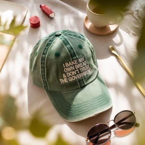 May include: A green baseball cap with the text "I BAKE MY OWN BREAD & DON'T TRUST THE GOVERNMENT" in white. The cap is on a white surface with sunglasses, a cup of tea, and other items.