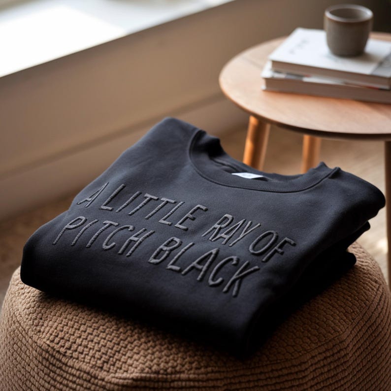 May include: A black sweatshirt with the text "A little ray of pitch black" embroidered on the front.