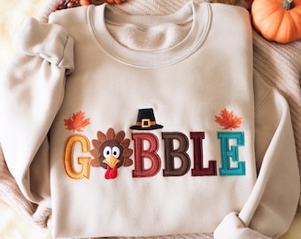 May include: Beige sweatshirt with the word "GOBBLE" embroidered in colorful letters. The "O" is a turkey with a red wattle, and the "B"s are topped with a pilgrim hat. Autumn leaves are on either side of the word.