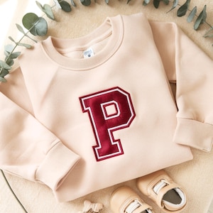 May include: A light peach-colored sweatshirt with a large, maroon letter "P" embroidered on the front. The letter is outlined in a darker red. The sweatshirt has a crew neck and long sleeves. A pair of baby shoes are visible.