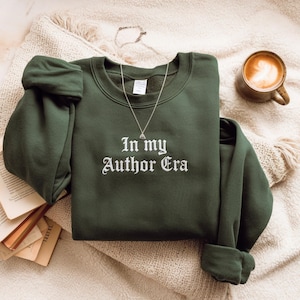 Könnte beinhalten: Ein dunkelgrünes Sweatshirt mit dem weißen Schriftzug "In my Author Era". Das Sweatshirt liegt gefaltet auf einer cremefarbenen Decke, mit einer silbernen Halskette und einer Kaffeetasse.