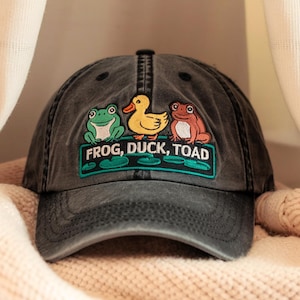 May include: A black, distressed, baseball cap with an embroidered patch featuring a green frog, a yellow duck, and a brown toad. The patch reads "Frog, Duck, Toad".