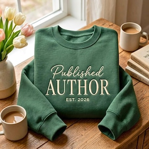 May include: A forest green crewneck sweatshirt featuring the cream-coloured text Published AUTHOR EST. 2026 centred on the chest. The garment has long sleeves with ribbed cuffs and a ribbed neckline, displayed on a wooden surface with books and flowers.