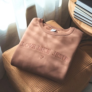 May include: A brown sweatshirt with the embroidered text "LOVE LIKE JESUS" and a heart symbol. The sweatshirt is folded neatly on a brown corduroy cushion. The image is well-lit, showcasing the details of the garment.