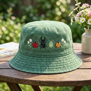 May include: A sage green bucket hat featuring embroidered details of a red ladybug, a black stag beetle, a light green beetle, and an orange ladybug, flanked by small white daisy flowers on both sides of the insect row on the front panel.