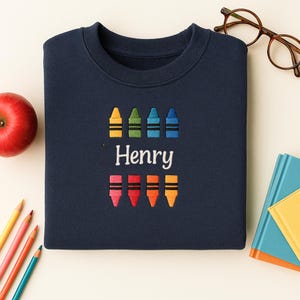 Op de afbeelding: Marineblauwe sweatshirt met geborduurde kleurpotloden in geel, groen, blauw en oranje. De naam "Henry" is onder de kleurpotloden geborduurd. Een rode appel, potloden en een bril zijn op de achtergrond te zien.