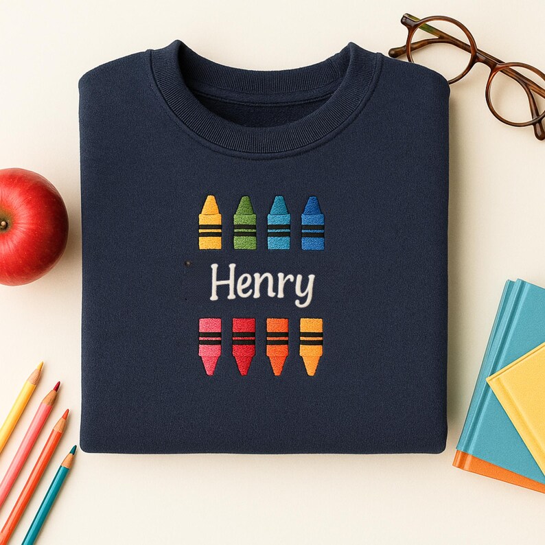 Op de afbeelding: Marineblauwe sweatshirt met geborduurde kleurpotloden in geel, groen, blauw en oranje. De naam "Henry" is onder de kleurpotloden geborduurd. Een rode appel, potloden en een bril zijn op de achtergrond te zien.
