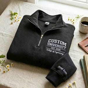 May include: Black quarter-zip sweatshirt with white embroidered text reading "CUSTOM UNIVERSITY EST. 2025 College of Engineering" on the chest. The sleeve cuff has "your icon" embroidered. The sweatshirt is on a neutral surface.