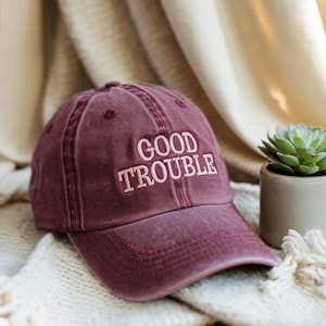 May include: A burgundy baseball cap with a white embroidered phrase "GOOD TROUBLE" on the front.