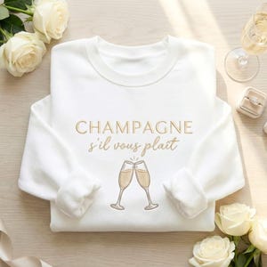 May include: White sweatshirt with the embroidered text "CHAMPAGNE s'il vous plaît" in gold. The design includes two champagne glasses clinking together. The image also features white roses, a ring box, and a champagne glass.