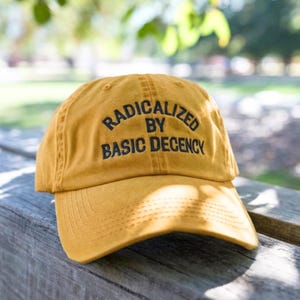 May include: A mustard yellow baseball cap featuring the embroidered black text RADICALIZED BY BASIC DECENCY across the front panels. The cap has a curved brim, a structured crown with stitched eyelets, and a classic six-panel construction.