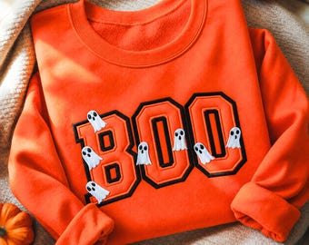 May include: A bright orange crewneck sweatshirt featuring the word "BOO" in large, block-style letters with a thick black outline. Several small, white, cartoon-style ghost figures with black eyes and mouths are layered over the letters.