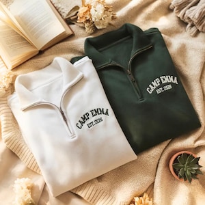 May include: A white and a dark green quarter-zip sweatshirt, each featuring the embroidered text CAMP EMMA EST. 2026 on the upper left chest. Both garments have a ribbed collar, a metal zipper, and long sleeves, presented in a folded arrangement.