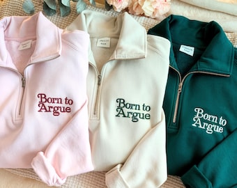 May include: Three quarter-zip sweatshirts in pastel pink, cream, and dark green. Each sweatshirt features the embroidered phrase "Born to Argue" in a contrasting color. The sweatshirts are displayed on a neutral-toned surface.