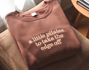 May include: A brown sweatshirt with the embroidered text "a little pilates to take the edge off." The crew neck sweatshirt is folded on a corduroy surface. The color of the sweatshirt is a muted brown.