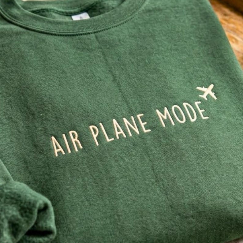 Puede incluir: Una sudadera verde oscuro con las palabras "AIR PLANE MODE" bordadas en color beige claro, junto con un peque&ntilde;o gr&aacute;fico de avi&oacute;n. La sudadera tiene cuello redondo y mangas largas.