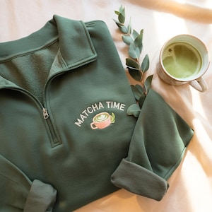 May include: Dark green sweatshirt with a quarter-zip neckline and embroidered "Matcha Time" design featuring a matcha latte. A cup of matcha tea and eucalyptus sprig are also in the image.
