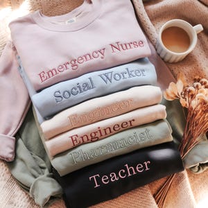 May include: A stack of six crewneck sweatshirts in various colors, each featuring embroidered text. The colors include light pink, light blue, beige, light peach, sage green, and black. The text reads Emergency Nurse, Social Worker, Engineer, Engineer, Pharmacist, and Teacher.