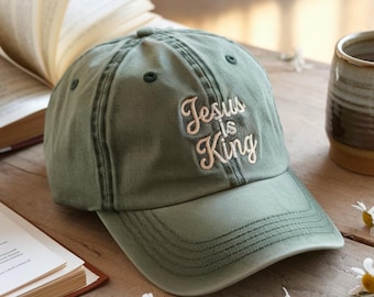 May include: An olive green baseball cap with the embroidered text "Jesus is King" in a cursive font. The cap has a curved brim and a classic six-panel design. The hat is made of a soft, worn-in fabric.
