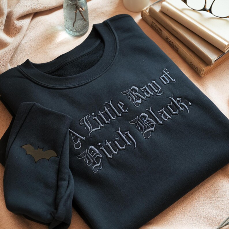 May include: Black sweatshirt and matching sweatpants set. The sweatshirt features the embroidered phrase "A Little Ray of Pitch Black." The sweatpants have a bat graphic on the cuff. The set is displayed on a pink surface.