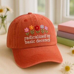 Könnte beinhalten: Eine rote Baseballkappe mit gestickten Blumen und dem Text "radikalisiert durch grundlegende Anständigkeit".