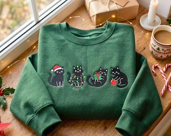 May include: A forest green crewneck sweatshirt featuring an embroidered graphic of four black cats. The first cat wears a red Santa hat, the second is tangled in colorful string lights, the third holds a green gift, and the fourth plays with a red ornament.
