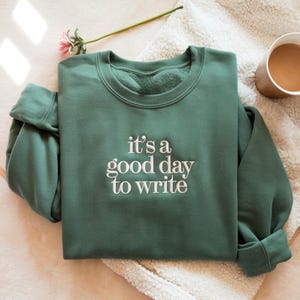 Könnte beinhalten: Ein dunkelgrünes Sweatshirt mit dem gestickten Schriftzug "it's a good day to write" in Weiß. Das Kleidungsstück ist auf einer weißen, strukturierten Oberfläche gefaltet, mit einer Tasse Kaffee und einer Blume im Hintergrund.