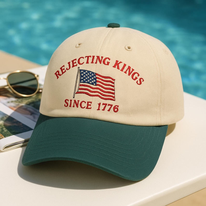 May include: A baseball cap with a cream-colored crown and a dark green brim. The cap features the text "REJECTING KINGS" and "SINCE 1776" embroidered in red, along with an American flag.