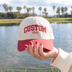 May include: A baseball cap with a cream-colored crown and a red brim. The word "CUSTOM" is embroidered in red on the front, with "TEXT" below it. The cap is held up in front of a lake.