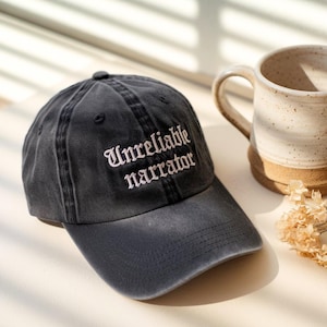 May include: A dark gray baseball cap with the embroidered text "Unreliable narrator" in white. The cap is slightly faded, with a curved brim and a small button on top. A ceramic mug is in the background.