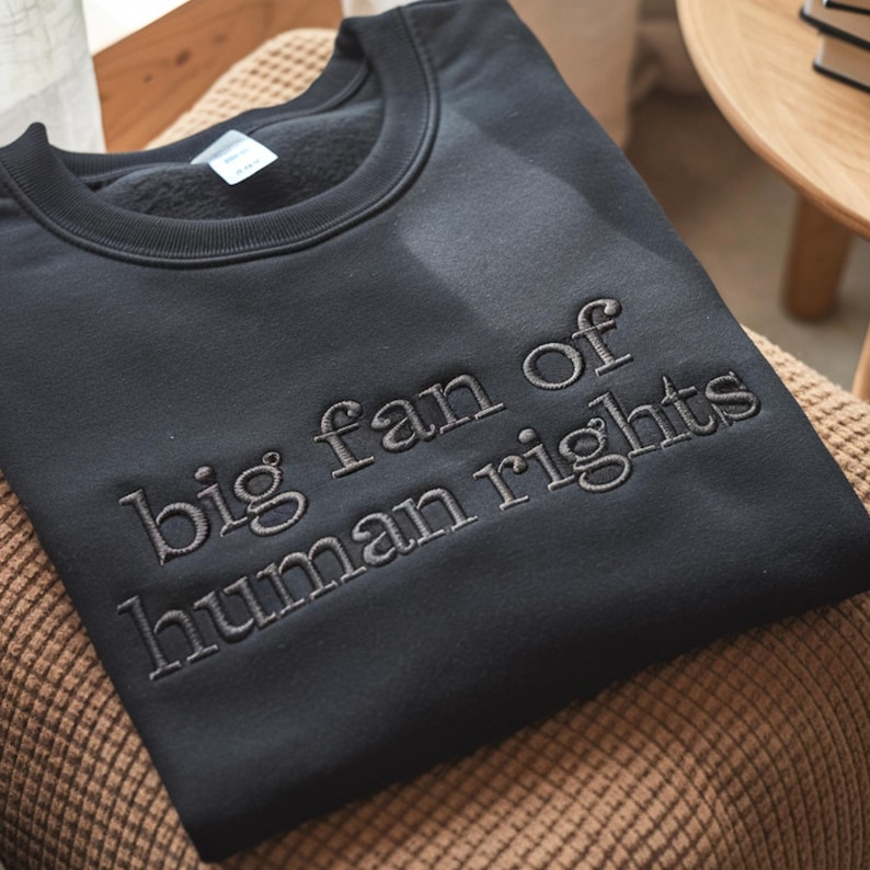 Puede incluir: Sudadera negra con la frase bordada "big fan of human rights" en gris claro. La sudadera de cuello redondo est&aacute; hecha de un tejido suave. La prenda se muestra sobre una silla.
