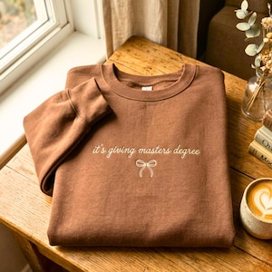 May include: A brown sweatshirt with the phrase "it's giving masters degree" embroidered in white, along with a bow graphic. The sweatshirt is folded on a wooden surface, with a coffee cup and books in the background.