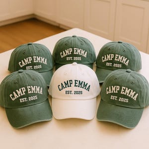 May include: Six baseball caps in a variety of colors. Five are a muted green, and one is white. Each cap has the words "CAMP EMMA" in a curved font, with "EST. 2025" below.