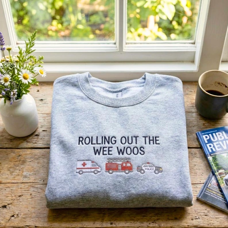 K&ouml;nnte beinhalten: Ein meliertes graues Sweatshirt mit dem gestickten Schriftzug "ROLLING OUT THE WEE WOOS" und Illustrationen eines Krankenwagens, eines Feuerwehrautos und eines Polizeiautos. Das Sweatshirt liegt gefaltet auf einem Holztisch.