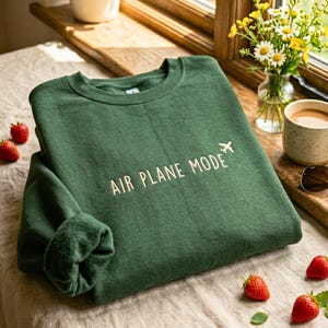 Puede incluir: Una sudadera verde oscuro con las palabras "AIR PLANE MODE" y un peque&ntilde;o avi&oacute;n blanco bordado. La sudadera est&aacute; doblada sobre una superficie clara, con fresas y una taza de caf&eacute; cerca.