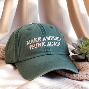Puede incluir: Una gorra de béisbol verde con el texto "Make America Think Again" bordado en blanco.